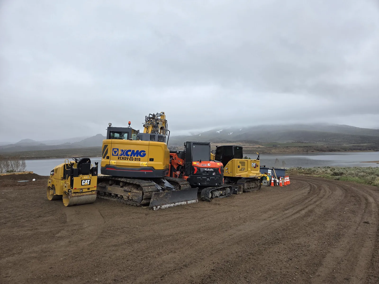 Excavators, a dozer and a roller lined up on a graded road by a lake